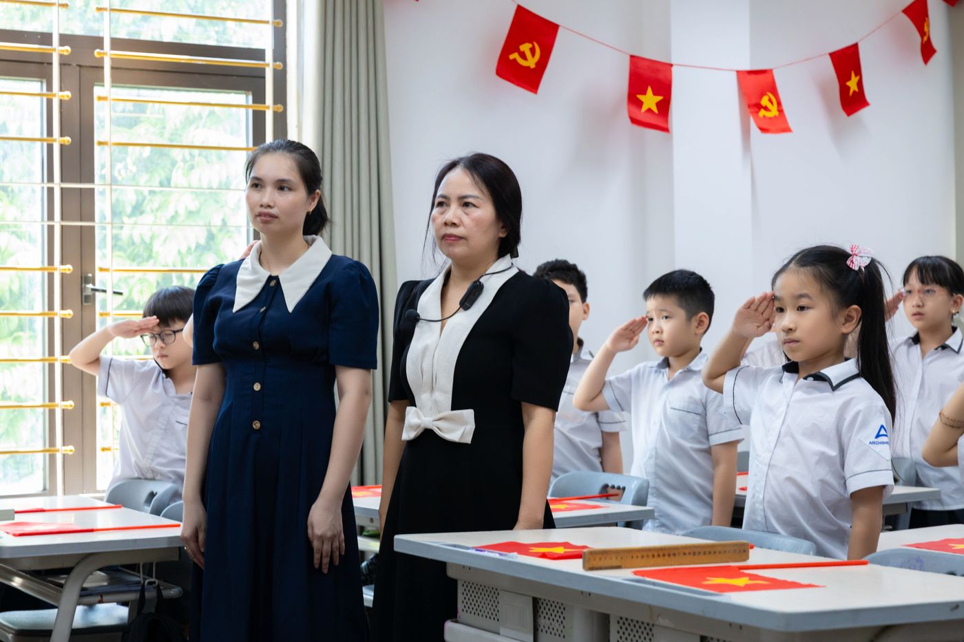 Hai cô giáo cùng học sinh trong giờ chào cờ tại một trường tiểu học